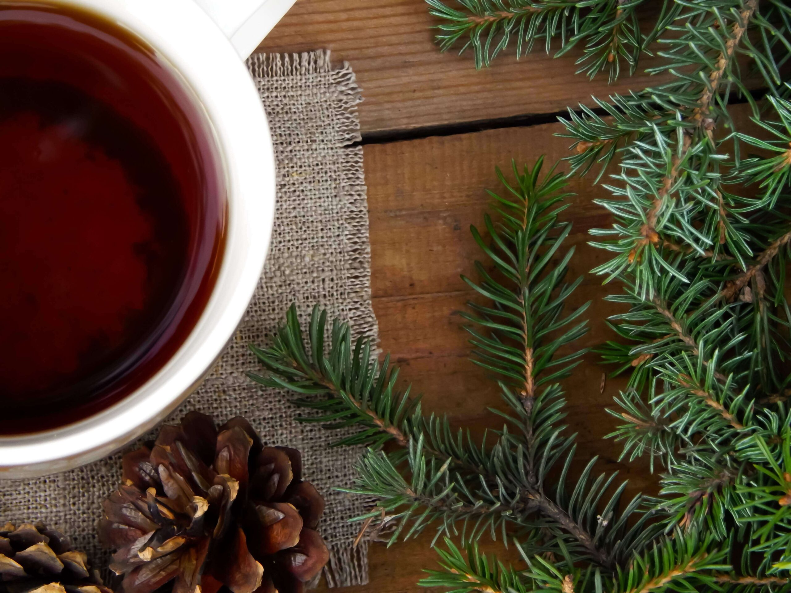 Tasse Tee auf Sackleinen mit Tannenzweigen und einem Tannenzapfen auf einer Holzoberfläche umgeben von Tannennadeln.