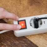 Eine Hand drückt den roten Netzschalter einer weißen Steckdosenleiste mit Steckern auf einer Holzoberfläche.