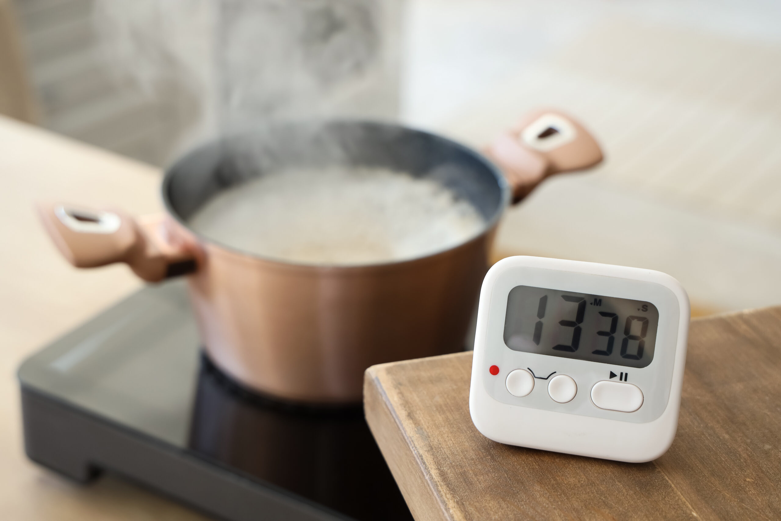 Auf einem Herd kocht ein Topf, auf einem Tisch in der Nähe steht ein digitaler Timer, der 13:38 anzeigt.