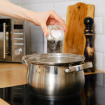 Eine Hand schüttet Salz in einen Topf mit Wasser auf einem Herd in einer modernen Küche.