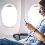 Frau sitzt am Flugzeugfenster, lächelt und benutzt ihr Smartphone während eines Fluges.