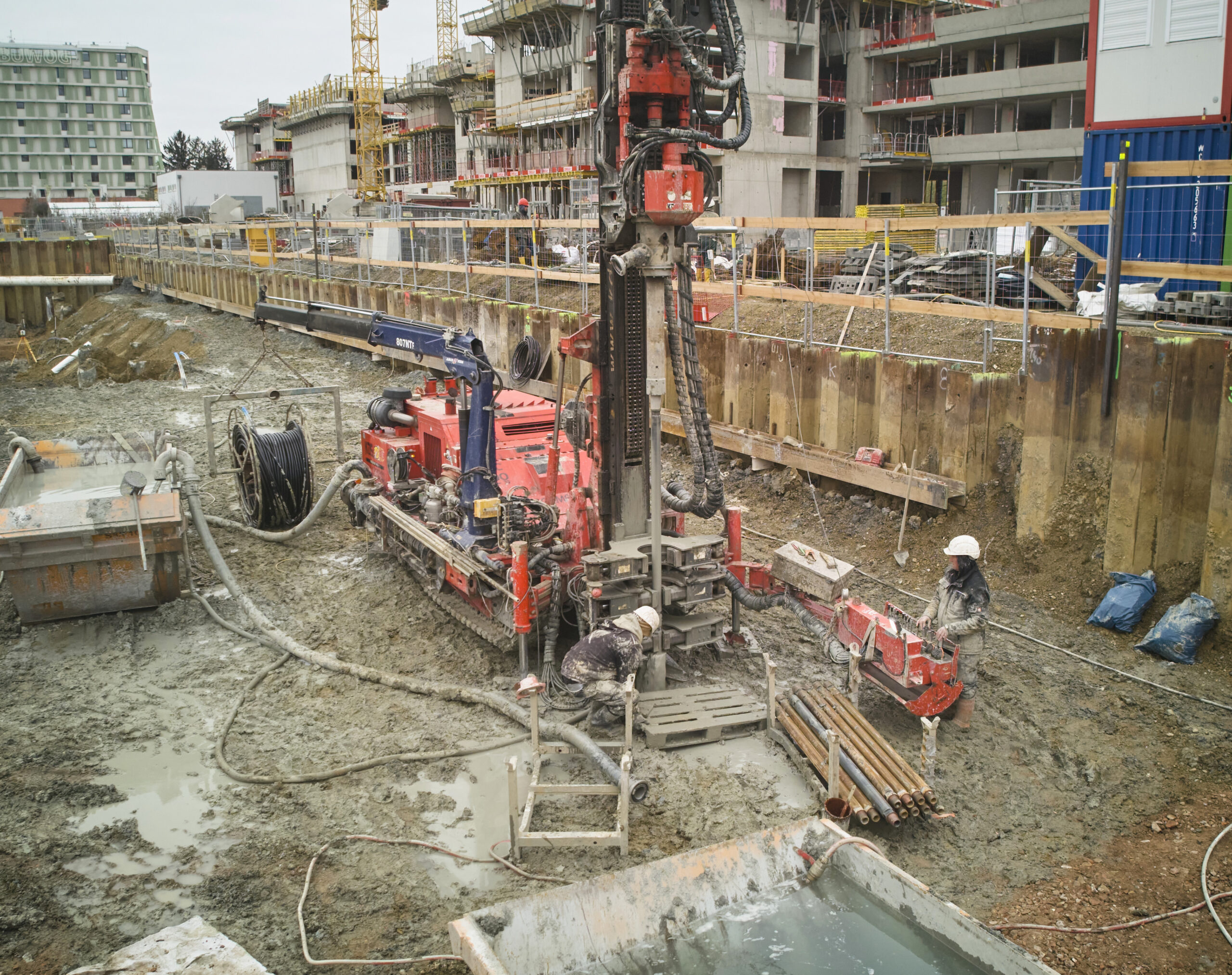 Eine rote Bohrmaschine auf einer schlammigen Baustelle bei der Installation von Tiefensonden, mit Arbeitern und Gebäuden im Hintergrund.