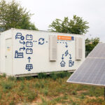 Sonnenkollektoren und weiße Energiespeicher mit blauen Schaltplänen, ausgestattet mit der innovativen „Second Life“-Batteriespeichertechnologie, im Freien aufgestellt, umgeben von Gras und Bäumen.