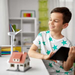 Lächelnder Junge, der ein Tablet in der Hand hält und an einem Schreibtisch mit einem Modell einer Windkraftanlage und einem Haus in einem hellen Raum sitzt.