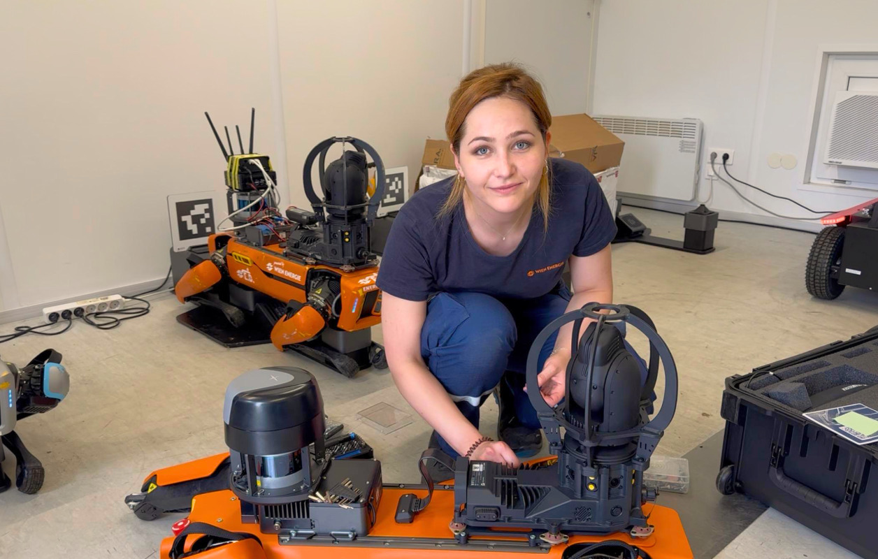 Charlotte Friedl kniet neben einem orangefarbenen Roboter in einem Raum, justiert dessen Komponenten und schaut in die Kamera.