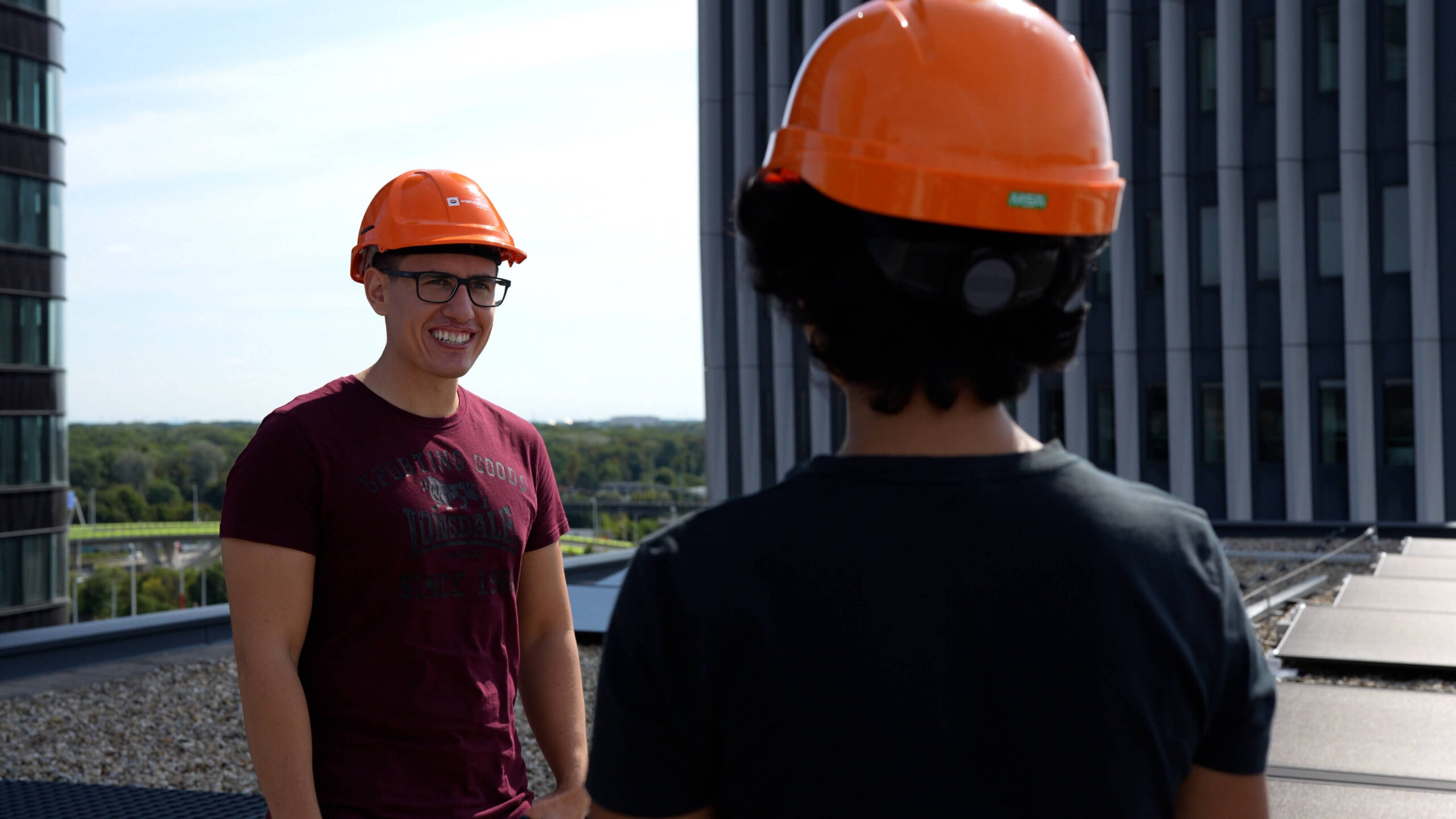Zwei Personen mit orangefarbenen Wien Energie Schutzhelmen unterhalten sich auf einem Dach, mit städtischen Gebäuden und Bäumen im Hintergrund, während Mario Mišković das Projekt beaufsichtigt.