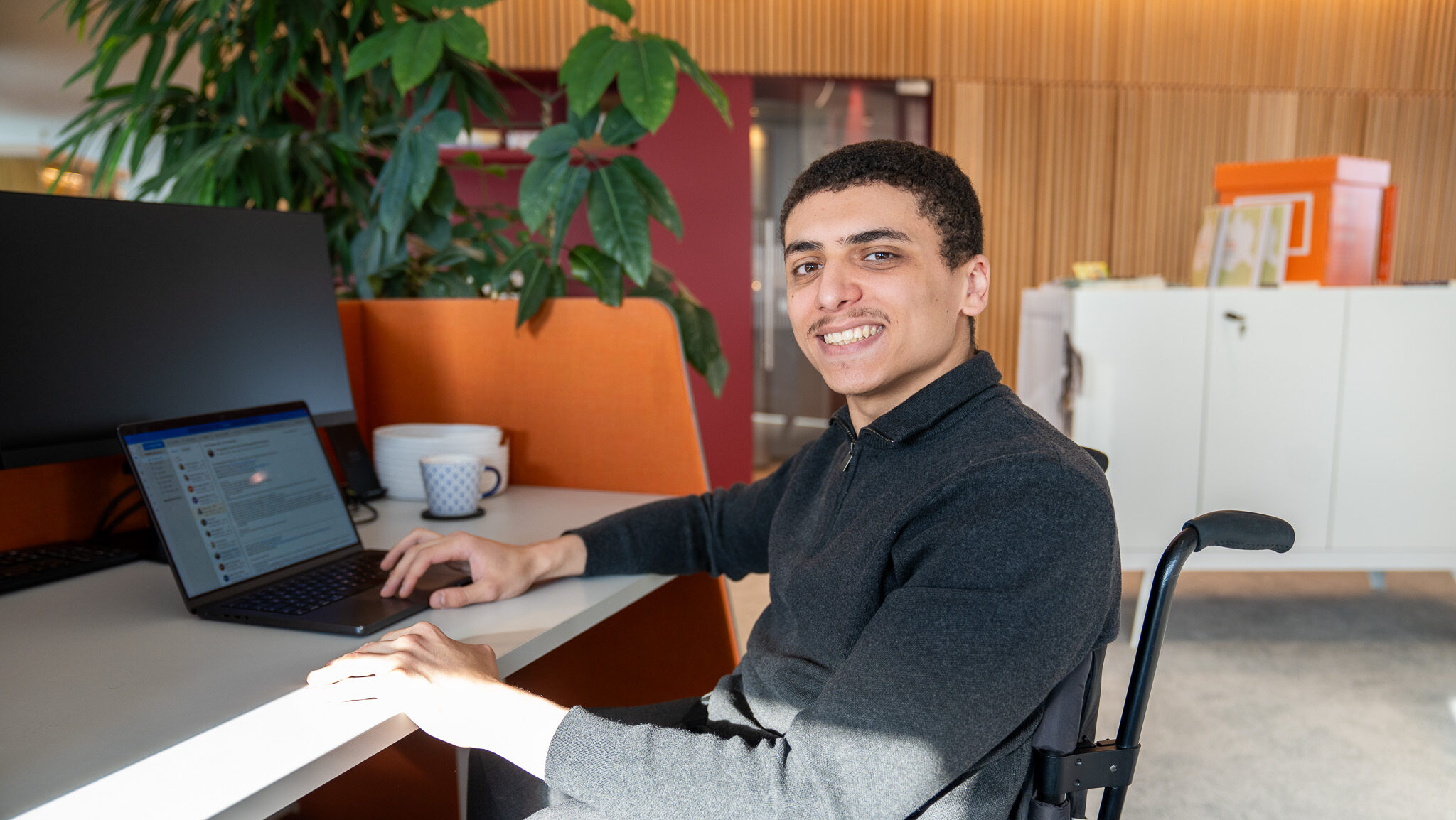 Nader-Abou-Abdou, ein Mann im Rollstuhl, lächelt und arbeitet an einem Laptop an einem Bürotisch.