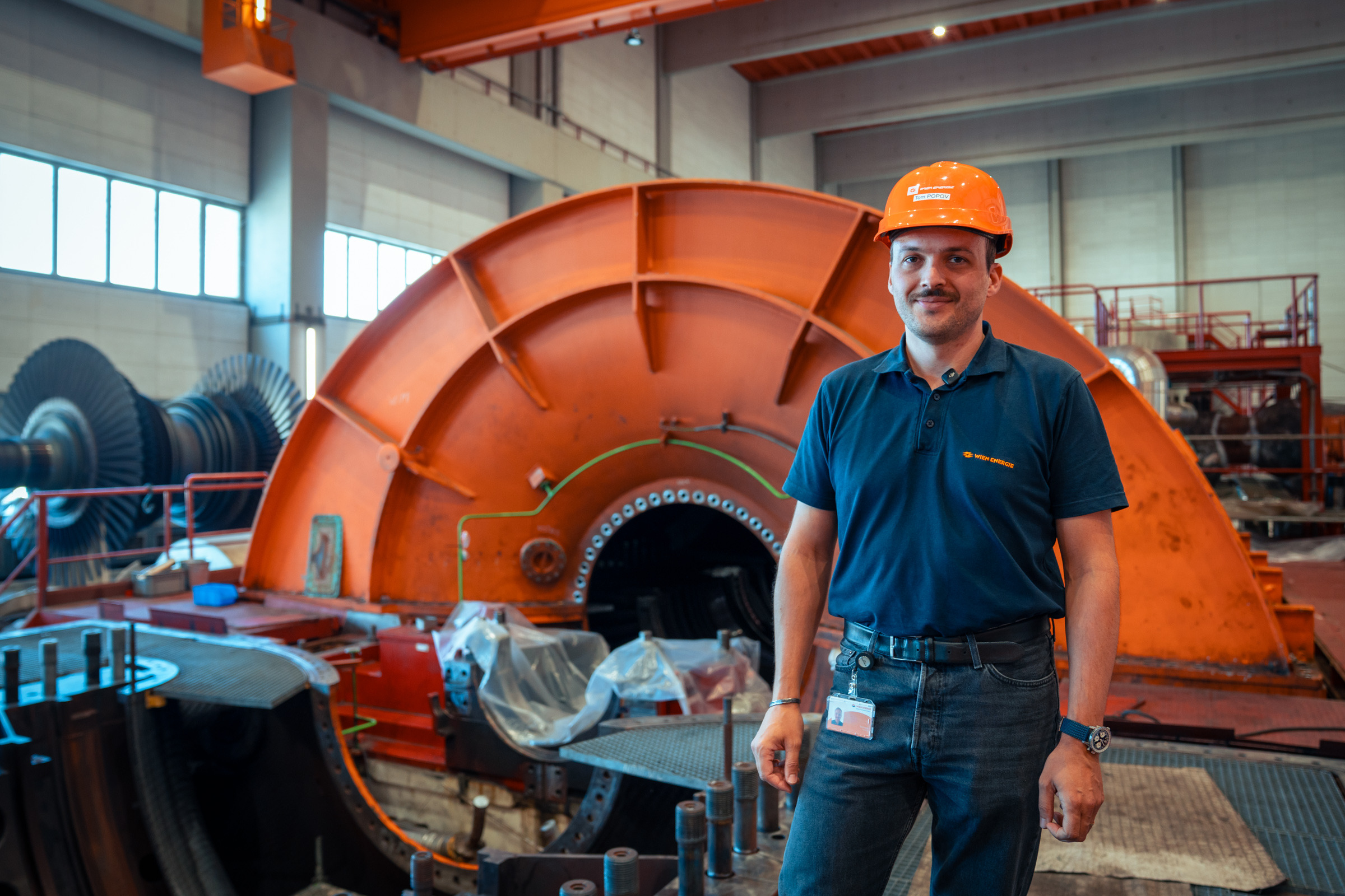 Tom Popov, der einen Schutzhelm trägt, steht vor großen Industriemaschinen in einer Fabrik oder einem Kraftwerk.
