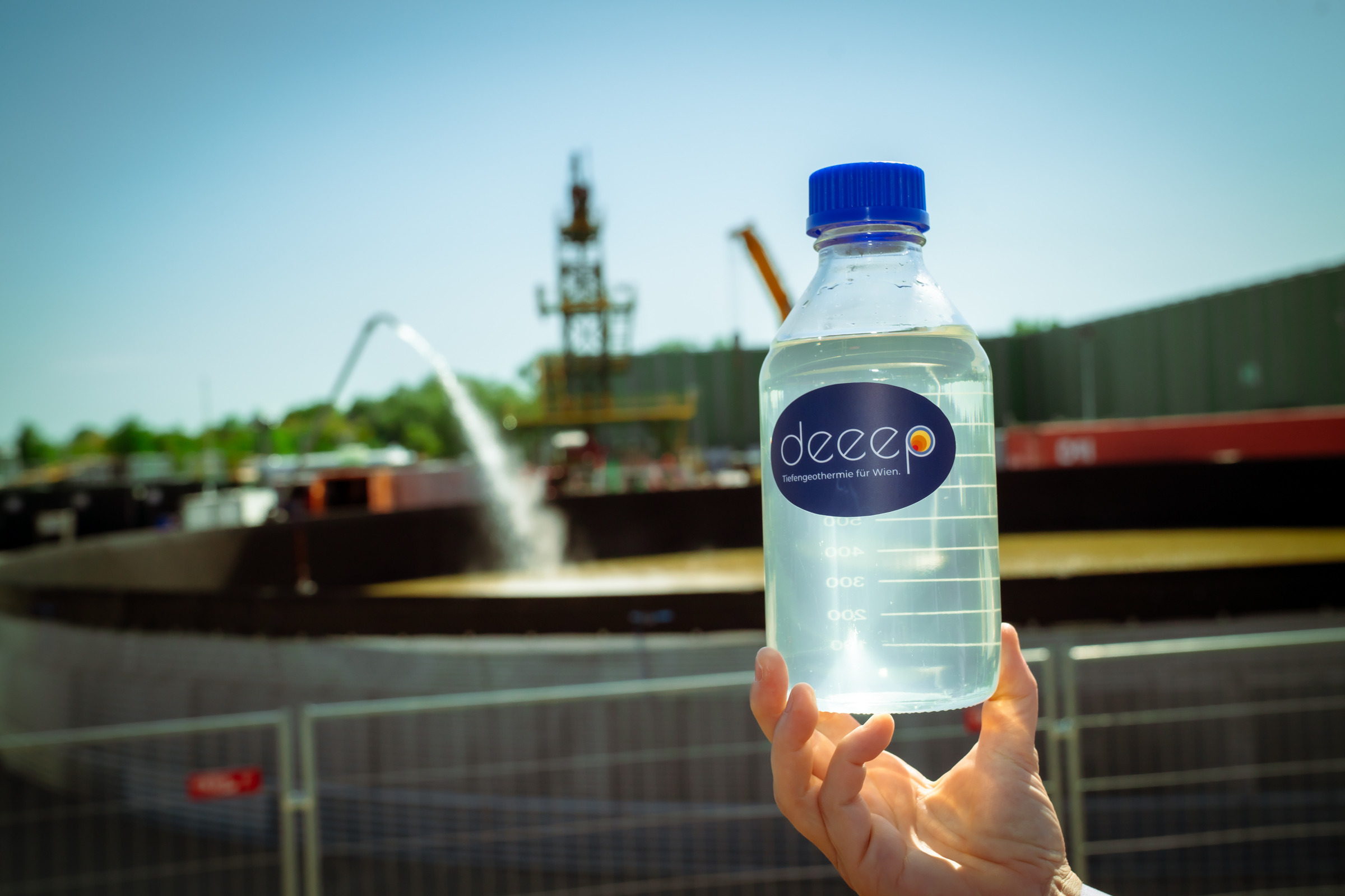 Eine Hand hält eine klare Wasserflasche mit der Aufschrift "deeep" vor der Tiefengeothermieanlage in Aspern, Wien.