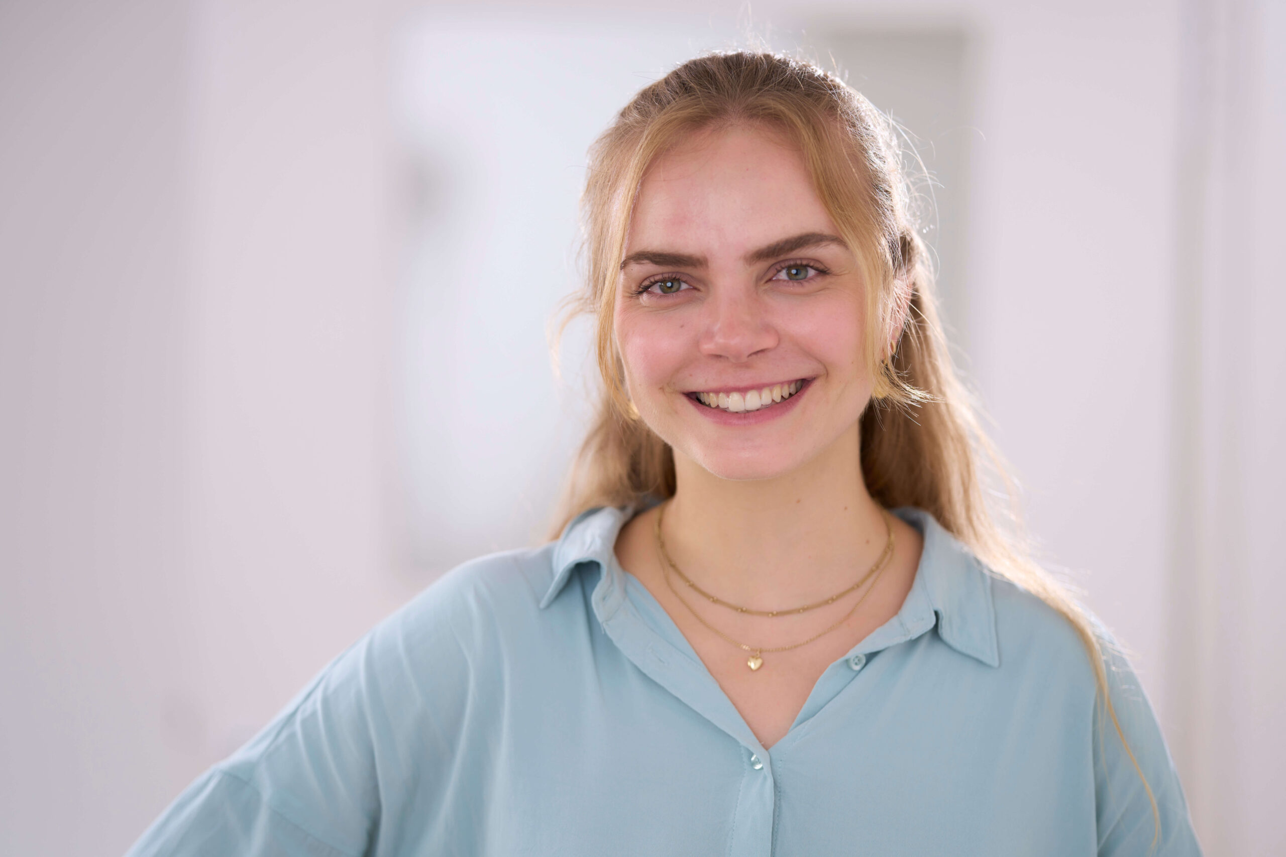 Leonie Machold, eine junge Frau mit langen blonden Haaren, lächelt, während sie ein hellblaues Hemd und goldene Halsketten trägt.