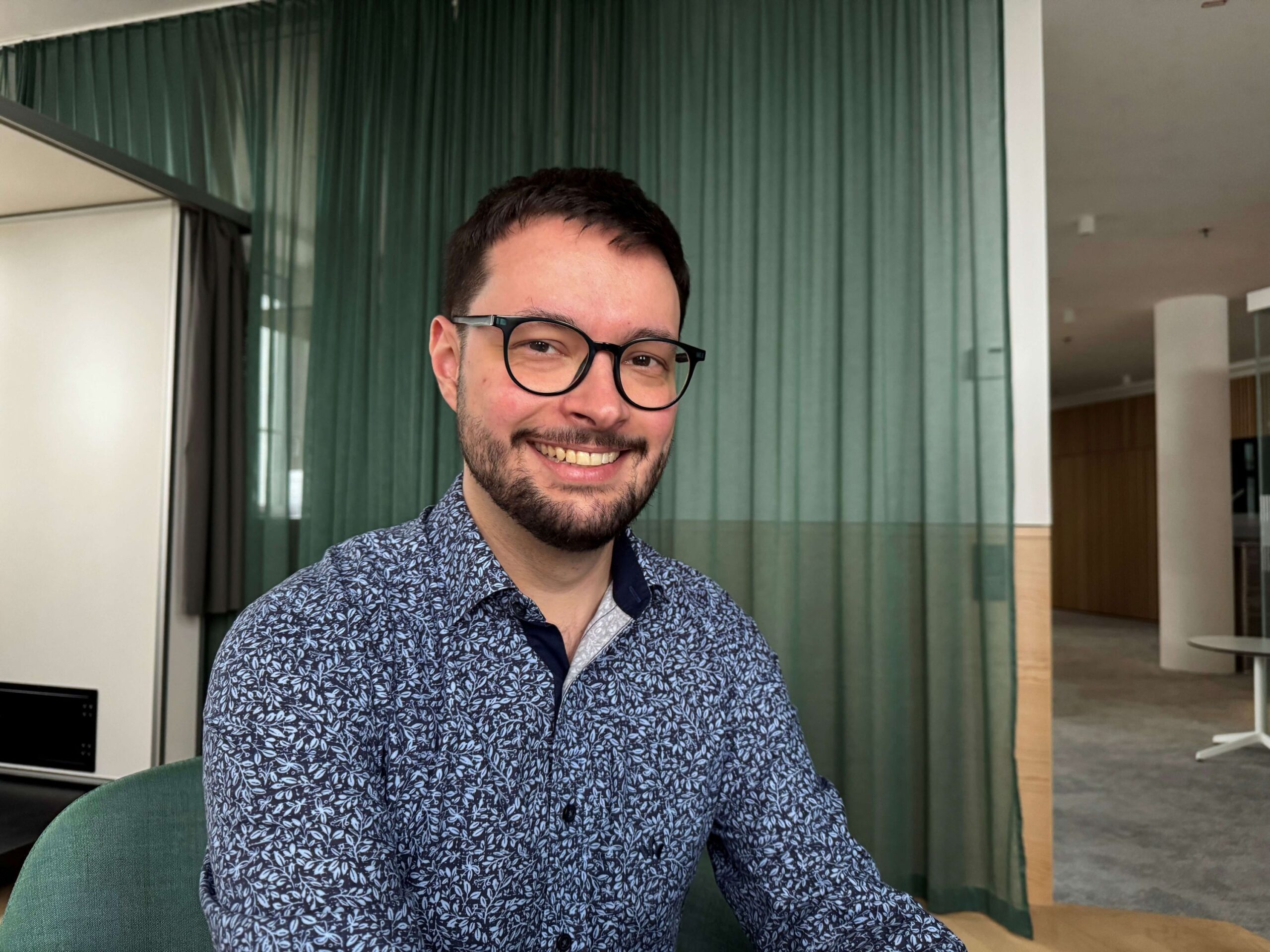 Manuel Harm, ein lächelnder Mann mit Brille und gemustertem Hemd, sitzt in einem Haus vor einem grünen Vorhang.