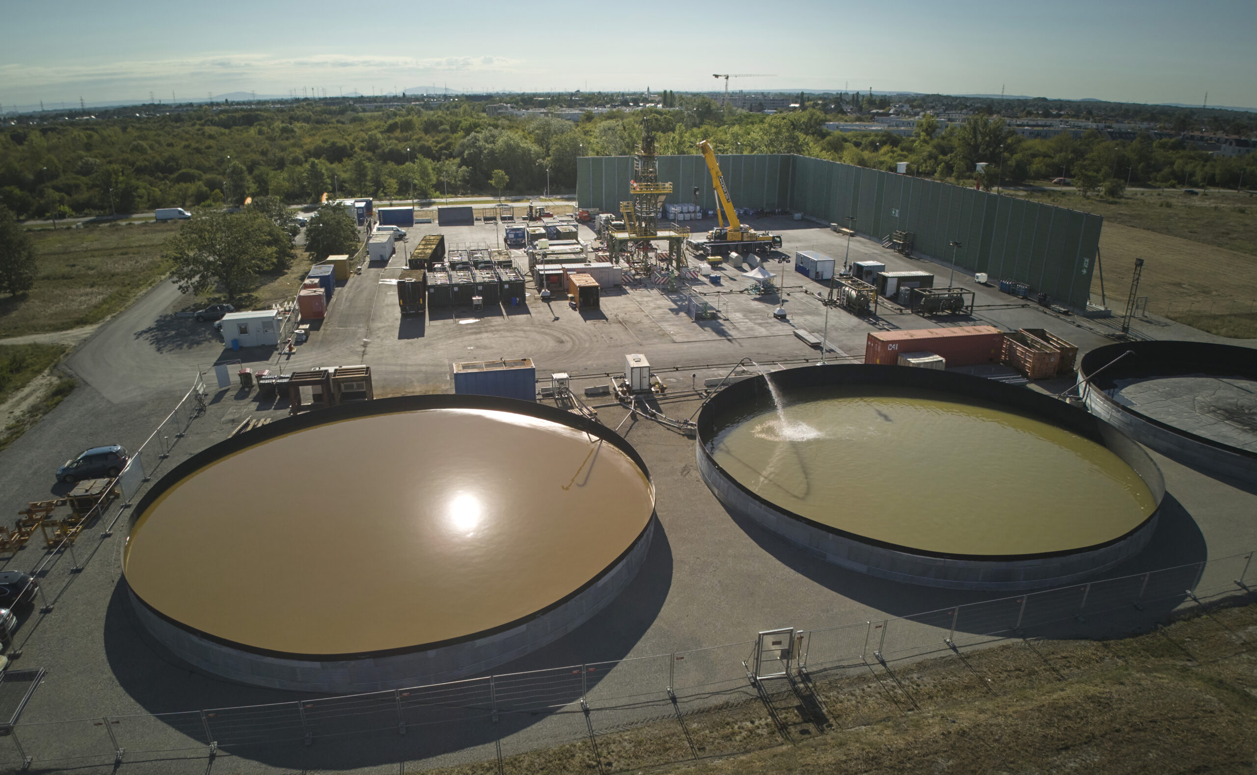 Luftaufnahme von zwei großen industriellen Wasserbecken beim Bohrplatz der Tiefengeothermie-Anlage in Aspern, umgeben von Geräten und Bäumen.