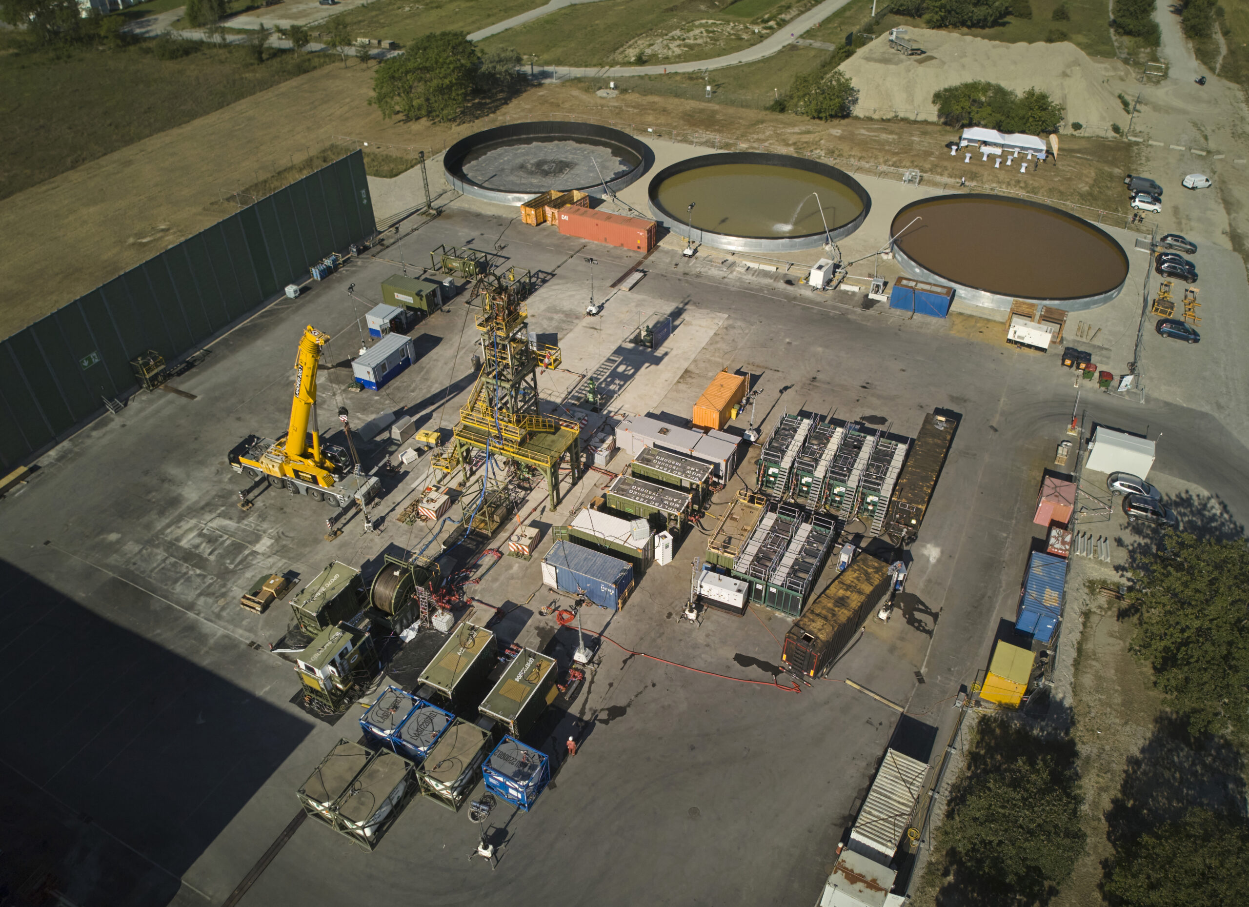 Luftaufnahme des Bohrplatzes für Wiens erste Tiefengeothermieanlage in Aspern mit in Reihen angeordneten Geräten, Fahrzeugen, Wasserbecken und Lagerbehältern.
