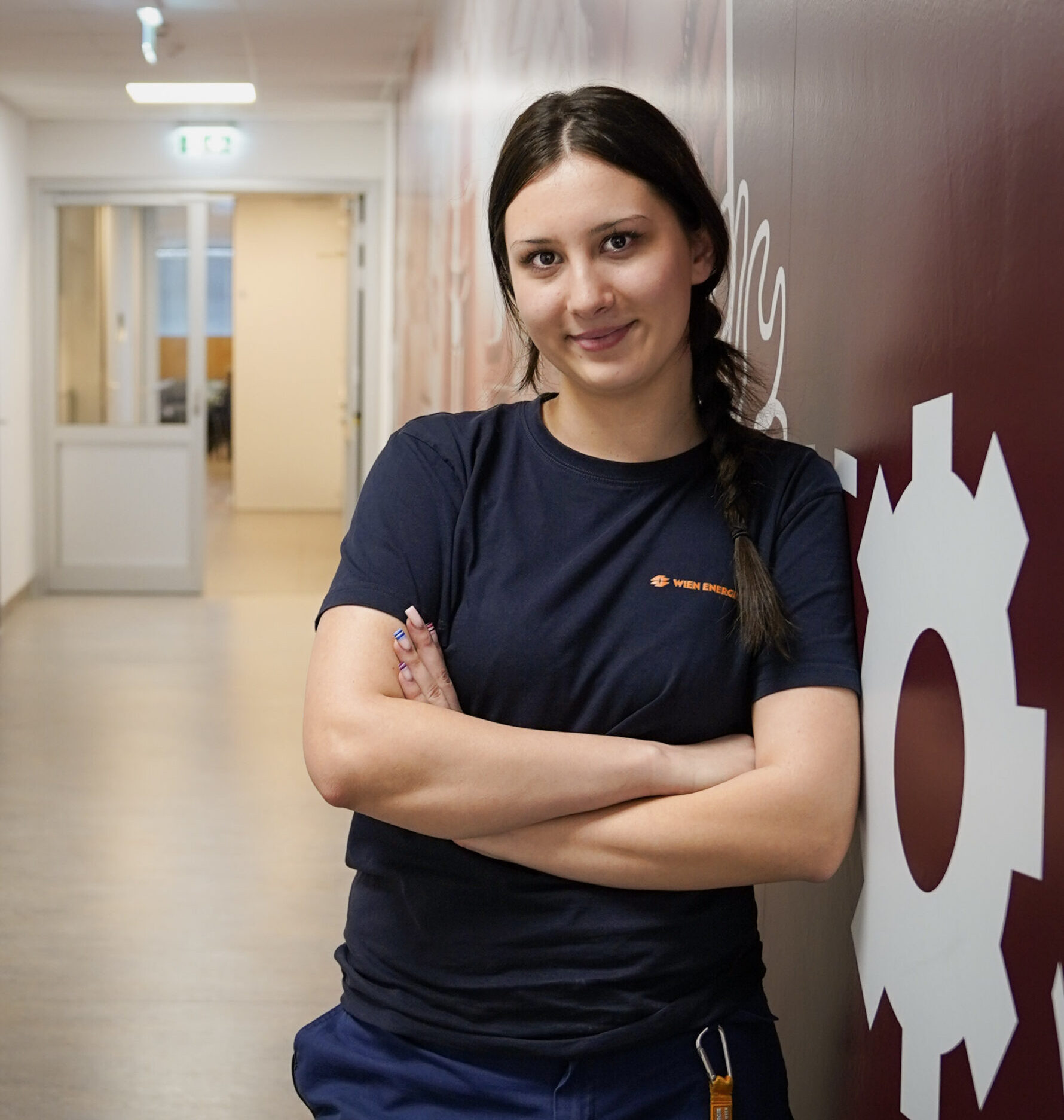 Nicole Menzel, mit geflochtenen Haaren, steht selbstbewusst mit verschränkten Armen an eine Wand mit einer großen Zahnradgrafik im Flur gelehnt.