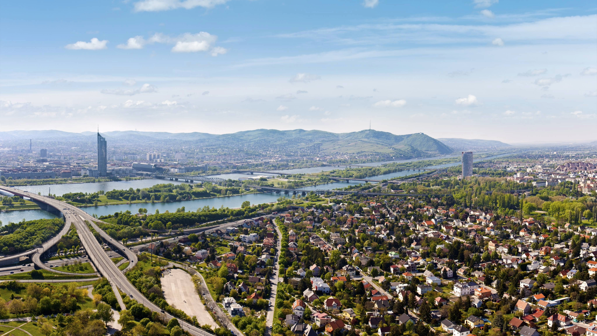 Luftaufnahme von Wien, auf der die Donau zu sehen ist, die sich durch die Stadt schlängelt, Brücken, die die Stadtteile miteinander verbinden, hoch in den Himmel ragende Gebäude und Hügel im Hintergrund unter einem strahlend blauen Himmel.