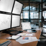 Vier Monitore und ein Laptop zeigen Aktiencharts auf einem Bürotisch mit Papieren, Kaffee und Geräten.