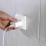 Eine Hand, die ein Handy-Ladegerät in eine weiße Steckdose an einer hellen Holzwand einsteckt.