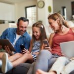 Eine vierköpfige Familie sitzt auf einem Sofa, wobei jeder ein Tablet oder Smartphone benutzt.