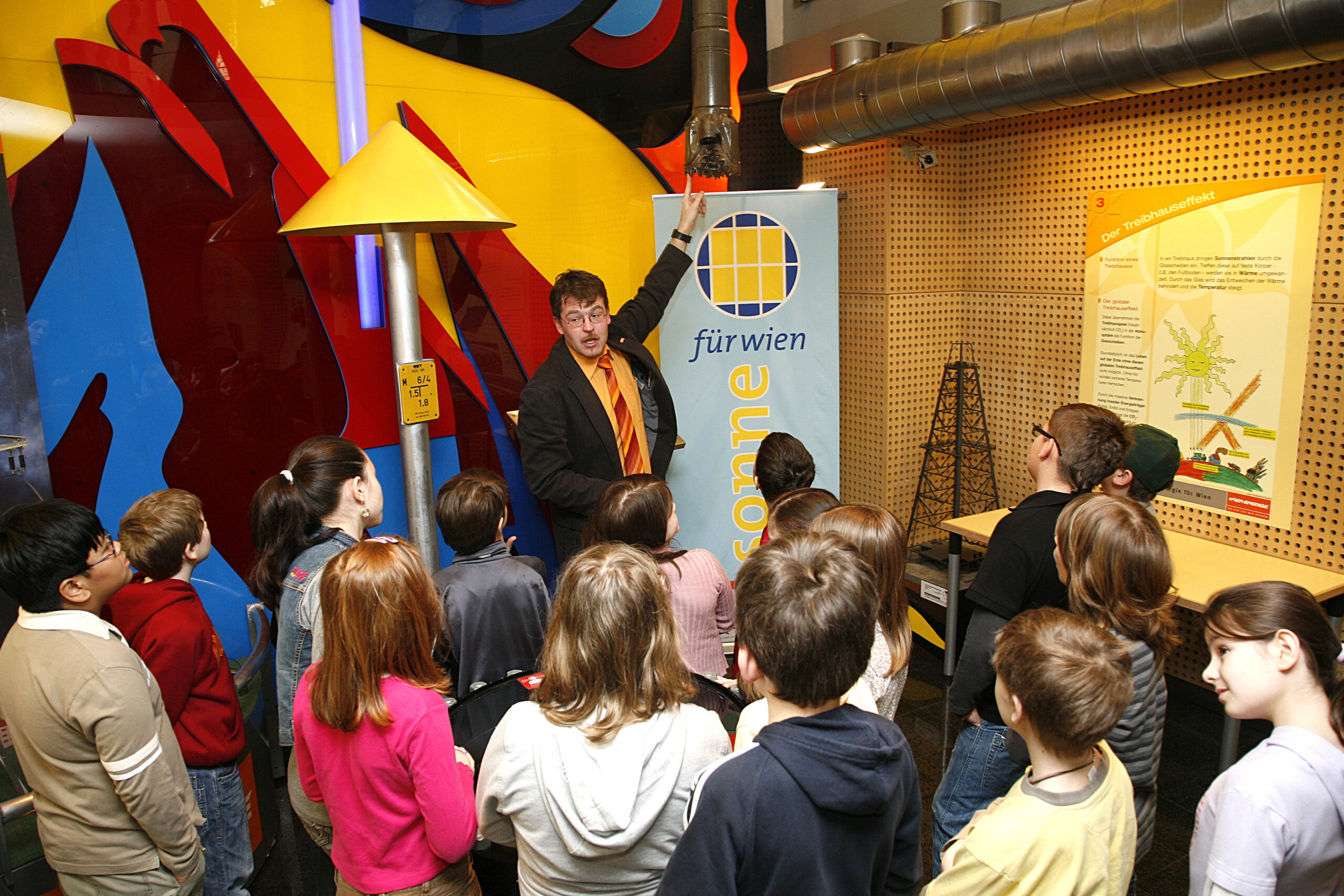 Ein Moderator spricht zu einer Gruppe von Kindern bei einer lehrreichen Ausstellung mit farbenfrohen Schaubildern im Wien Energie Haus.