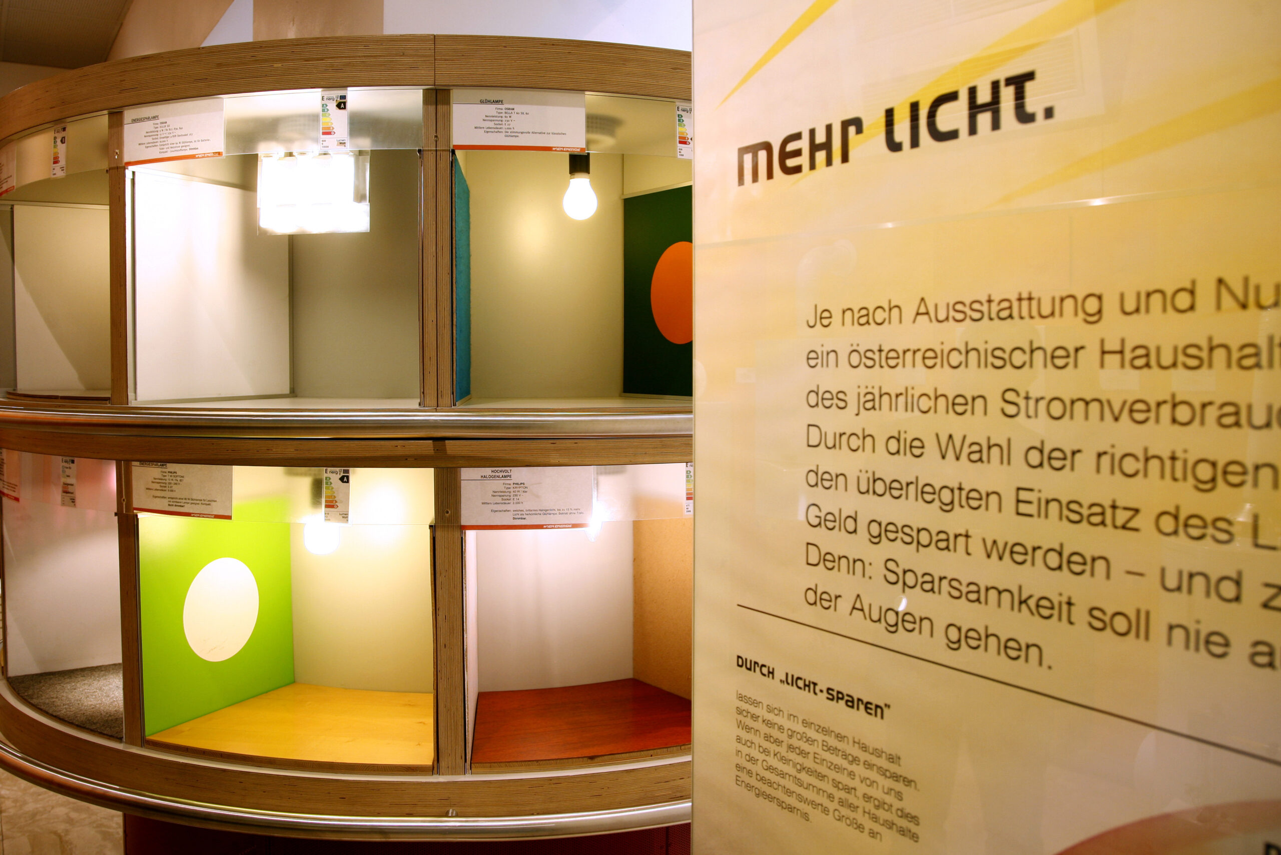 Anzeige von Energiesparlampen in verschiedenfarbigen Kästen mit dem deutschen Text „mehr Licht“ auf einer vertikalen Tafel.