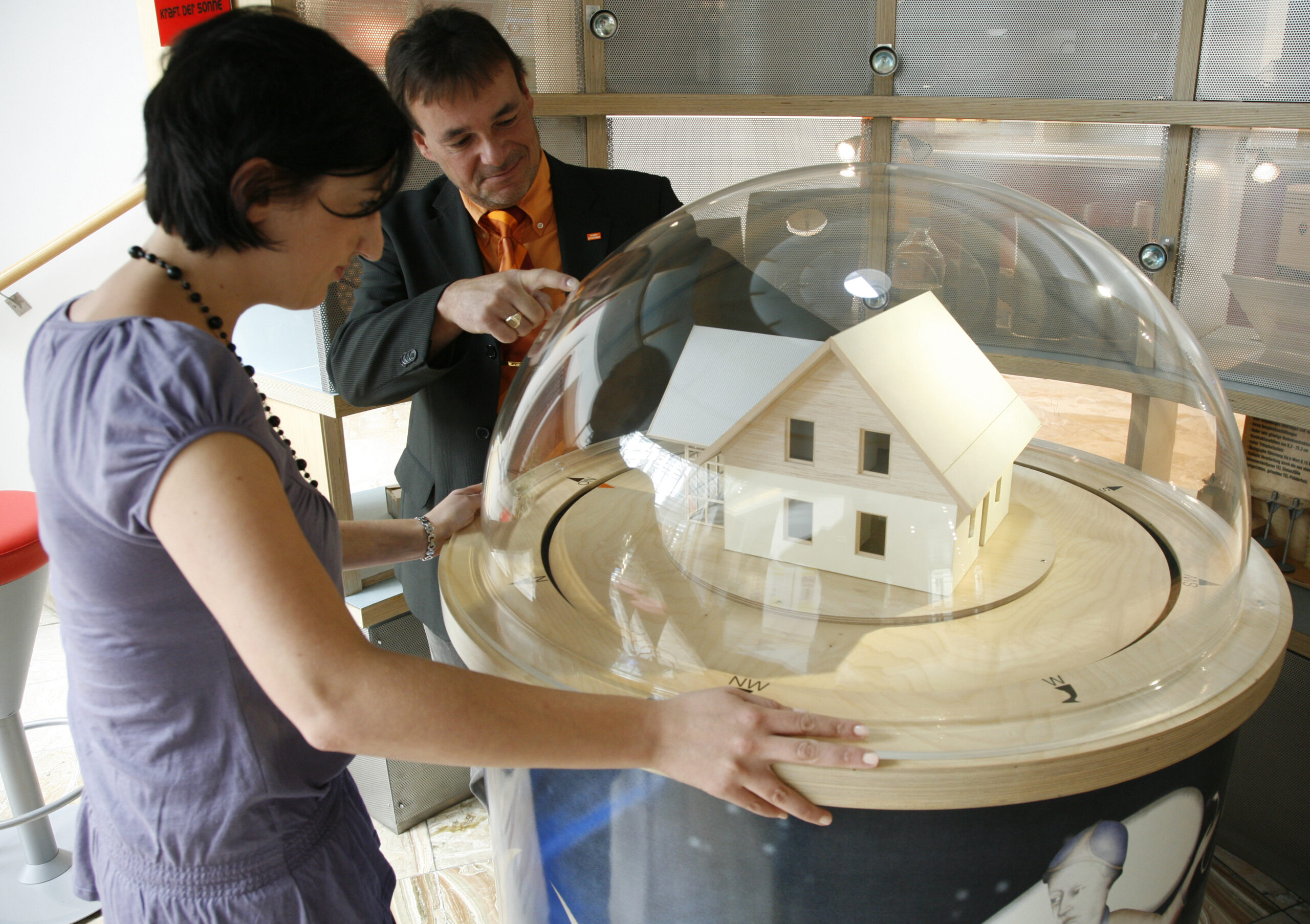 Zwei Personen untersuchen ein Modellhaus in einer durchsichtigen Kuppel. Eine Person zeigt mit dem Finger darauf, während die andere genau hinsieht.